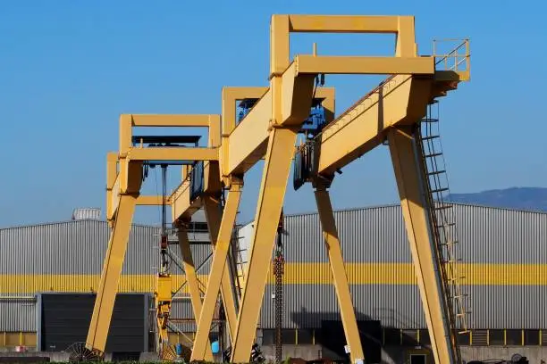 Gantry cranes in front of a gray and yellow building of a manufacturing factory.