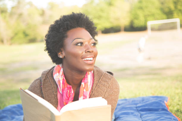 One African descent woman outdoors reading a book in a park during spring or summer season.  The beautiful young adult wears a pink shirt and jacket and lies on a blue blanket.