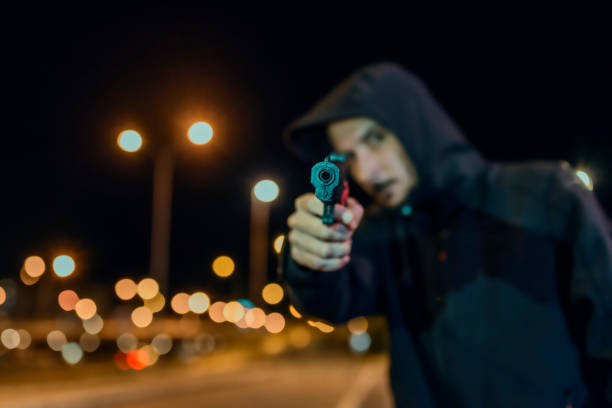 Using deadly force Photo of Robber in black hoodie with a gun, concept of danger, threat and extortion. Man in the dark pointing a firearm at his victim against a night street background, looking at camera with serious face. man holding gun at night in the city stock pictures, royalty-free photos & images