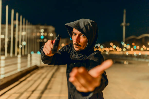 Your money or your life Photo of Robber in black hoodie with a gun holds his hand out for the money, concept of danger, threat and extortion. Man in the dark pointing a firearm at his victim against a night street background, looking at camera with serious face. man holding gun at night in the city stock pictures, royalty-free photos & images