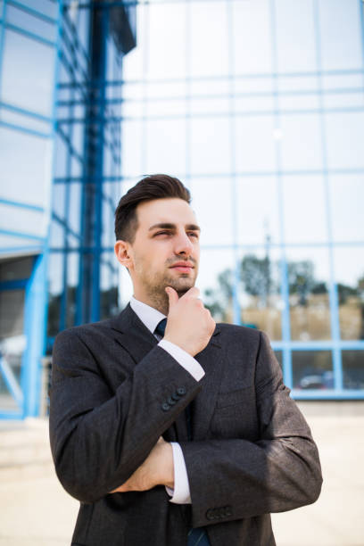 istock Confident thoughtful businessman in a suit holding hand on chin and looking away while standing outdoors with office building in the background 862011052