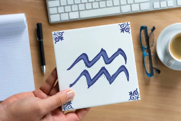 Person holding Delfts Blue aquarius zodiac tile above working desk. Drawing on white tile by contributor