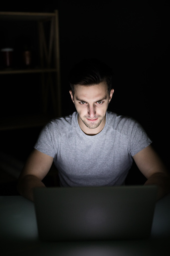 Handsome man reading news on laptop lying in dark room. Looking concentrated