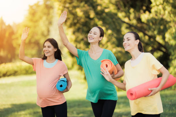 three pregnant girls posing in a park with yoga mats in hand. they smile and have fun - human pregnancy outdoors women nature imagens e fotografias de stock