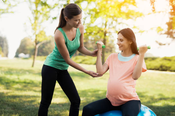 pregnancy yoga. a coach helps a pregnant woman to do exercises on a ball for yoga - human pregnancy outdoors women nature imagens e fotografias de stock