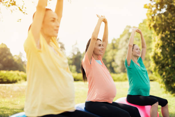 three pregnant women are sitting in a park on yoga balls with their hands up - human pregnancy outdoors women nature imagens e fotografias de stock