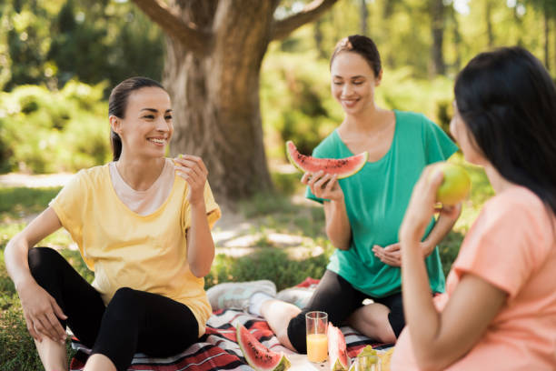 three pregnant women sit in the park on a rug for picnics and eat. they are all smiling - human pregnancy outdoors women nature imagens e fotografias de stock