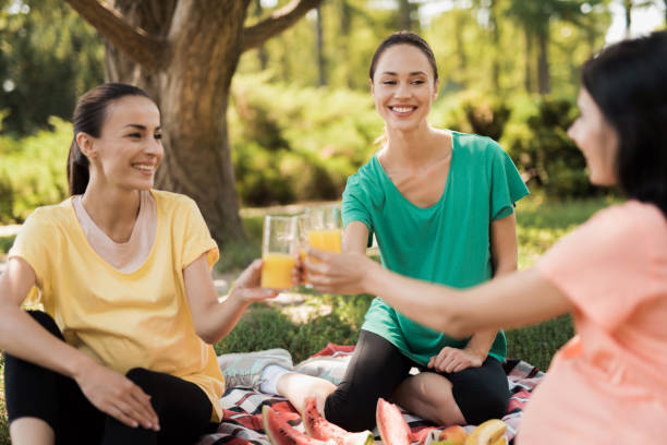 three pregnant women sit in the park on a rug for picnics and eat. they are all smiling. they clink glasses with juice - human pregnancy outdoors women nature imagens e fotografias de stock