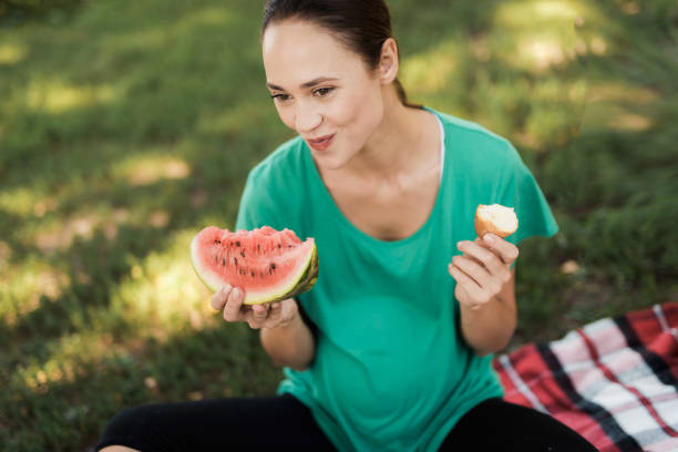 pregnant woman in a green t-shirt sits on a picnic in the park and eats watermelon - child women outdoors mother imagens e fotografias de stock