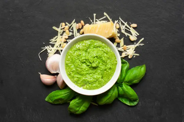Pesto sauce in a bowl with pine nuts, parmesan and garlic over black stone background. top view