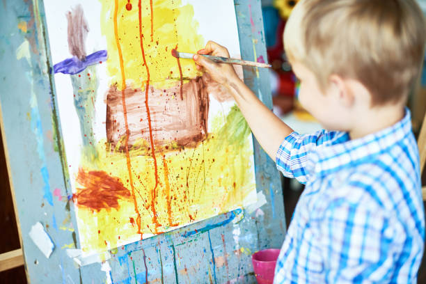 Back view portrait of little boy painting picture of house using watercolors on easel in art studio