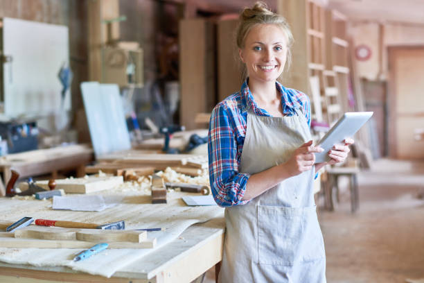Young Carpenter Using Digital Tablet in Woodworks Shop Portrait of happy young woman looking at camera using digital tablet in modern woodworking shop, copy space carpenter carpentry craftsperson carving stock pictures, royalty-free photos & images