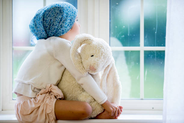 Sitting At The Windowsill A young Ethnic girl is indoors in a hospital room. She has cancer. She is wearing casual clothing and a bandanna to hide her hair loss. She is sitting and staring out the window while holding a stuffed animal. hospital depression sadness bed stock pictures, royalty-free photos & images