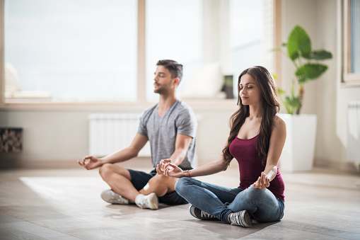 Zen-like couple practicing Yoga with eyes closed while sitting on the floor in empty room of their new home. Focus is on woman.