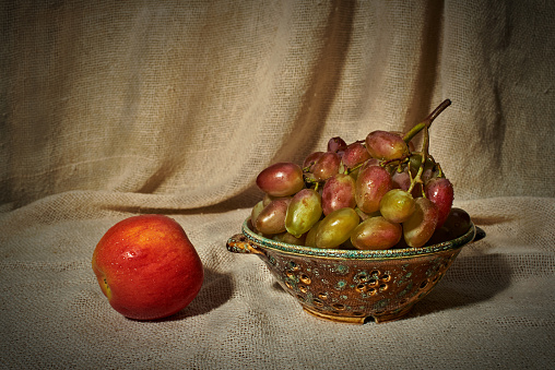 Still life with an apple, grapes and a colander on the background of a curtain of coarse cloth. On fruits are drops of water
