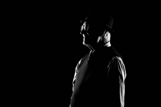 Side view of old-fashioned senior man in hat and glasses looking straight in dark room