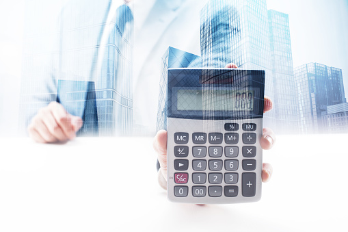 Businessman sitting by the desk holding a calculator with double exposure cityscape.