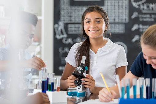 Pretty Hispanic female middle school student uses a microscope while working on science assignment with her friends.