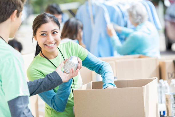 Male and female volunteers sort donations during food drive Male and female teenage food bank volunteers sort canned food items in cardboard boxes. caregivers day stock pictures, royalty-free photos & images