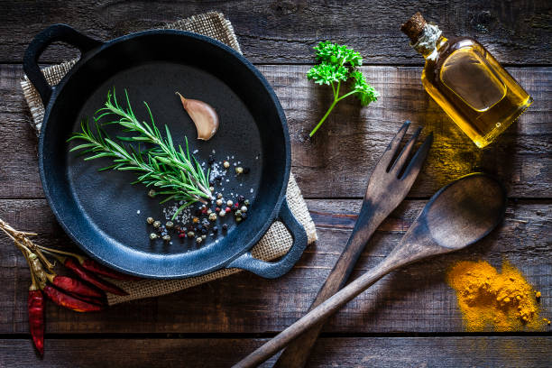 cucina: padella in ghisa nera con spezie ed erbe aromatiche sul tavolo da cucina in legno - spice condiment spoon wooden spoon foto e immagini stock