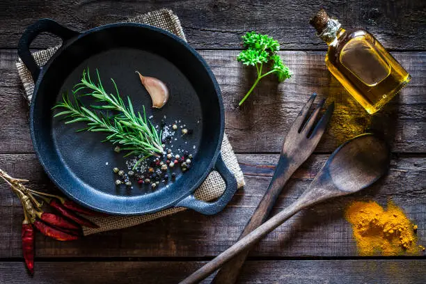 Top view of a black cast iron pan with some spices and herbs like rosemary, parsley, garlic clove, peppercorns, salt, olive oil, chili pepper and turmeric shot on rustic wooden kitchen table. A burlap tissue is under the cooking pan and an old wooden spoon and fork are beside it. Low key DSRL studio photo taken with Canon EOS 5D Mk II and Canon EF 100mm f/2.8L Macro IS USM