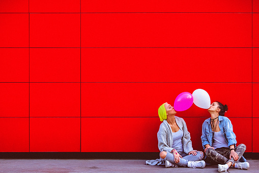 Two young female friends having fun, blowing up balloons in front of the red wall