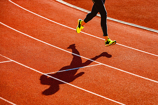 Sportsperson running over the orange running track. Sports clothing - sports shoes and running tights. High angle view of a runner's legs and its deep shadow on the track.
