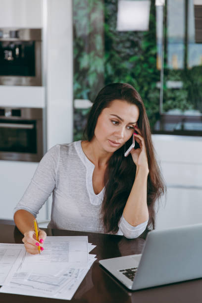 istock Attractive young modern business woman talking on mobile phone and working with documents and laptop in the kitchen at home 862431290