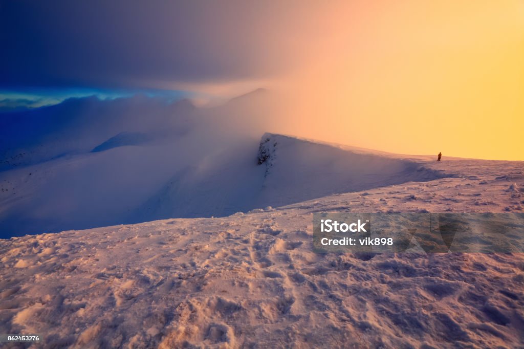At the edge of the snowy rock someone is standing. The high mountains in the fog, morning sky is enlighten with the orange colored light in winter day. Adventure Stock Photo