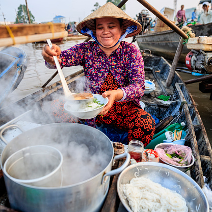 Vietnamese woman selling famous pho bo -noodle soup, floating market on Mekong River Delta, South Vietnam. Pho bo is a extremely popular Vietnamese noodle soup consisting of broth, rice noodles, herbs and beef meat.