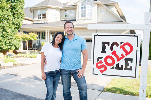 A Caucasian husband and wife are posing outdoors in front of a new house. They are wearing casual clothing. They are standing beside the sold sign and smiling at the camera while embracing.