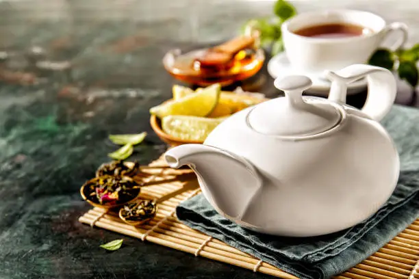 Arranged white ceramic teapot with various tea addition on black surface.