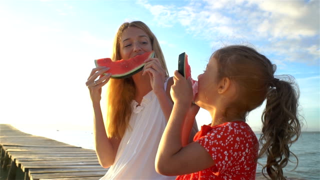 Happy mother and her daughter sitting on a wooden pier and eating a juicy watermelon