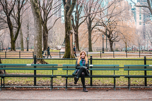 Photo of Girl sitting on a bench in front of trees at the Central Park in Manhattan, New York City