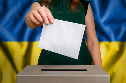 Election in Ukraine - voting at the ballot box. The hand of woman putting her vote in the ballot box. Flag of Ukraine on background.
