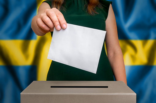 Election in Sweden - voting at the ballot box. The hand of woman putting her vote in the ballot box. Flag of Sweden on background.