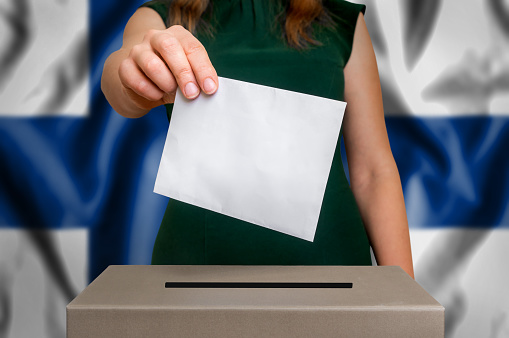 Election in Finland - voting at the ballot box. The hand of woman putting her vote in the ballot box. Flag of Finland on background.