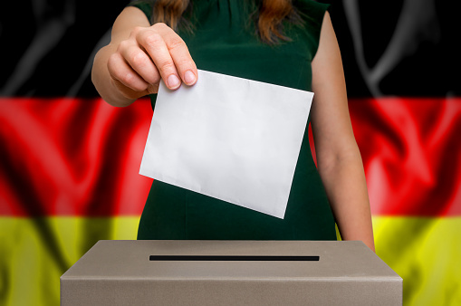 Election in Germany - voting at the ballot box. The hand of woman putting her vote in the ballot box. Flag of Germany on background.