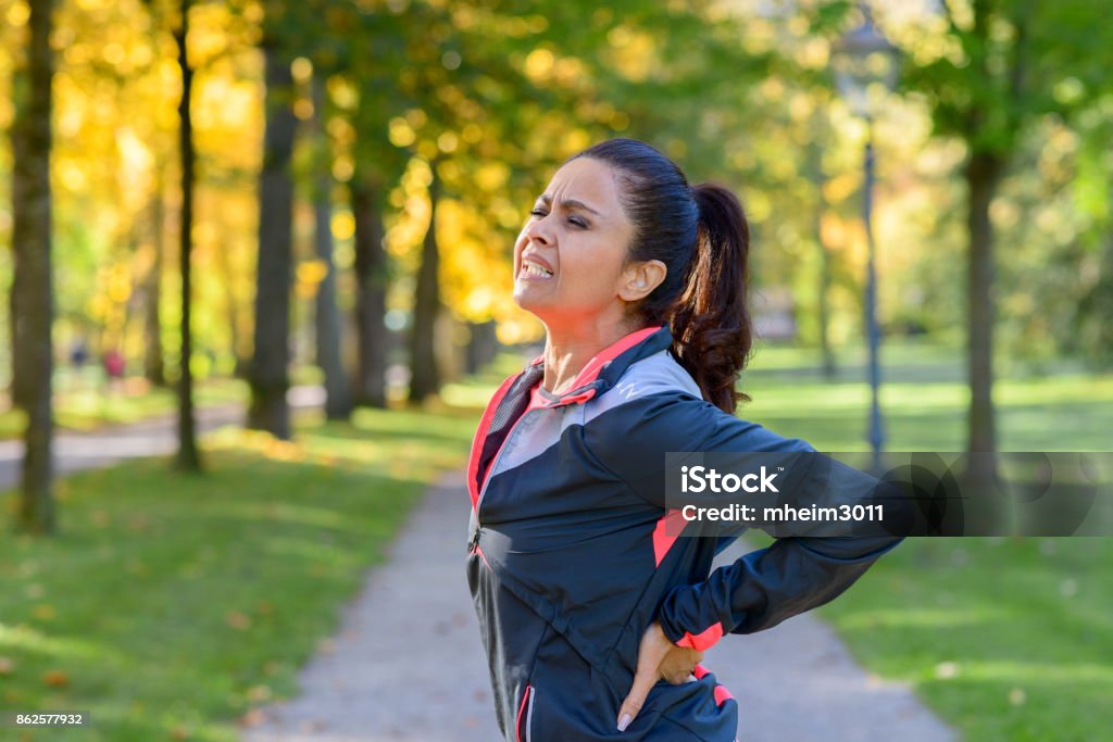 Woman holding painful back in park Adult woman holding her painful back after exercising in park Backache Stock Photo