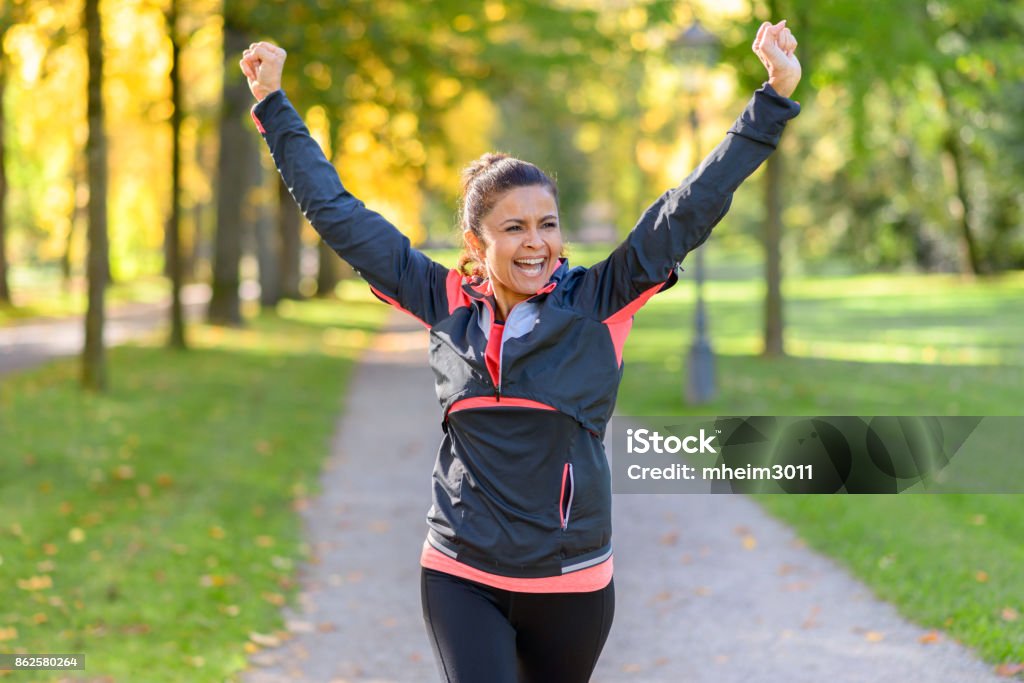 Happy fit woman cheering and celebrating Happy fit middle aged woman cheering and celebrating as she walks along a rural lane through a leafy green park after working out jogging Exercising Stock Photo