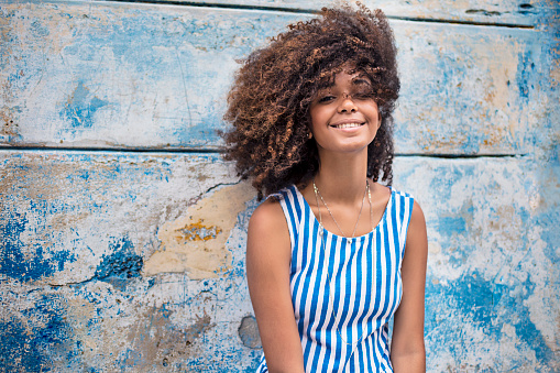 Smiling young female is standing against weathered blue wall. Portrait of beautiful woman is with Afro hairstyle. She is wearing striped sleeveless dress.