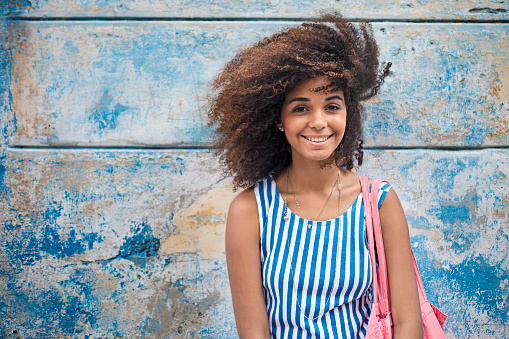 Portrait of beautiful woman with Afro hairstyle. Smiling young female is standing against weathered blue wall. She is wearing striped sleeveless dress.