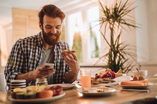 Young smiling man eating a sandwich at dining table and typing on his smart phone.