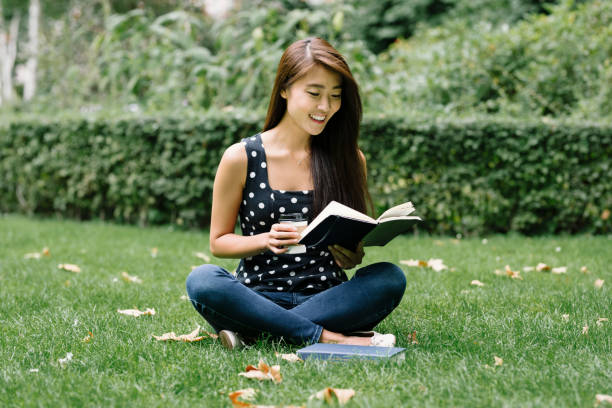 Young Asian student studying in a public park in London. She has taken her books and coffee with her to stay fresh and focused during her studies.