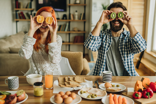 Young happy couple covering their eyes with fresh fruit and having fun at dining table.