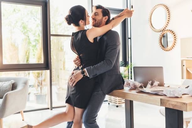 Young couple embracing in office Young man in suit sitting on desk in office hugging woman in black dress and leaning in for kiss work romance stock pictures, royalty-free photos & images
