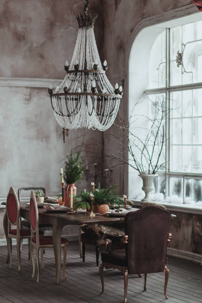 istock Modern loft dining room with high ceiling, vintage armchairs, empty grey concrete wall, wooden floor, dining table with dishes, flowers and candles. Big crystal chandelier hanging from the ceiling. Mock up cozy interior photo 862657296