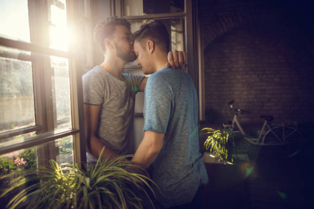Unconditional love in homosexual relationship! Loving gay men standing by the window while one of them is kissing his boyfriend in the forehead. man gay stock pictures, royalty-free photos & images