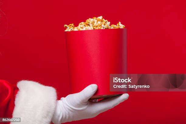 Christmas Photo Of Santa Claus Gloved Hand With A Red Bucket With Popcorn On A Red Background Stock Photo - Download Image Now