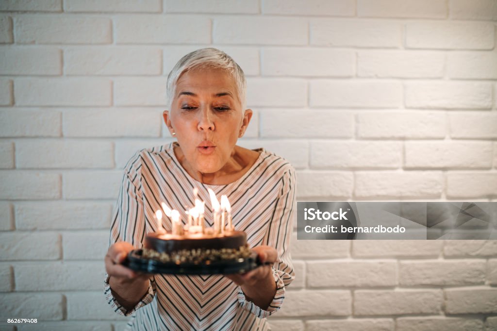 Make a wish! A beautiful senior lady making a wish and blowing candles on her lovely birthday cake. Blowing Stock Photo